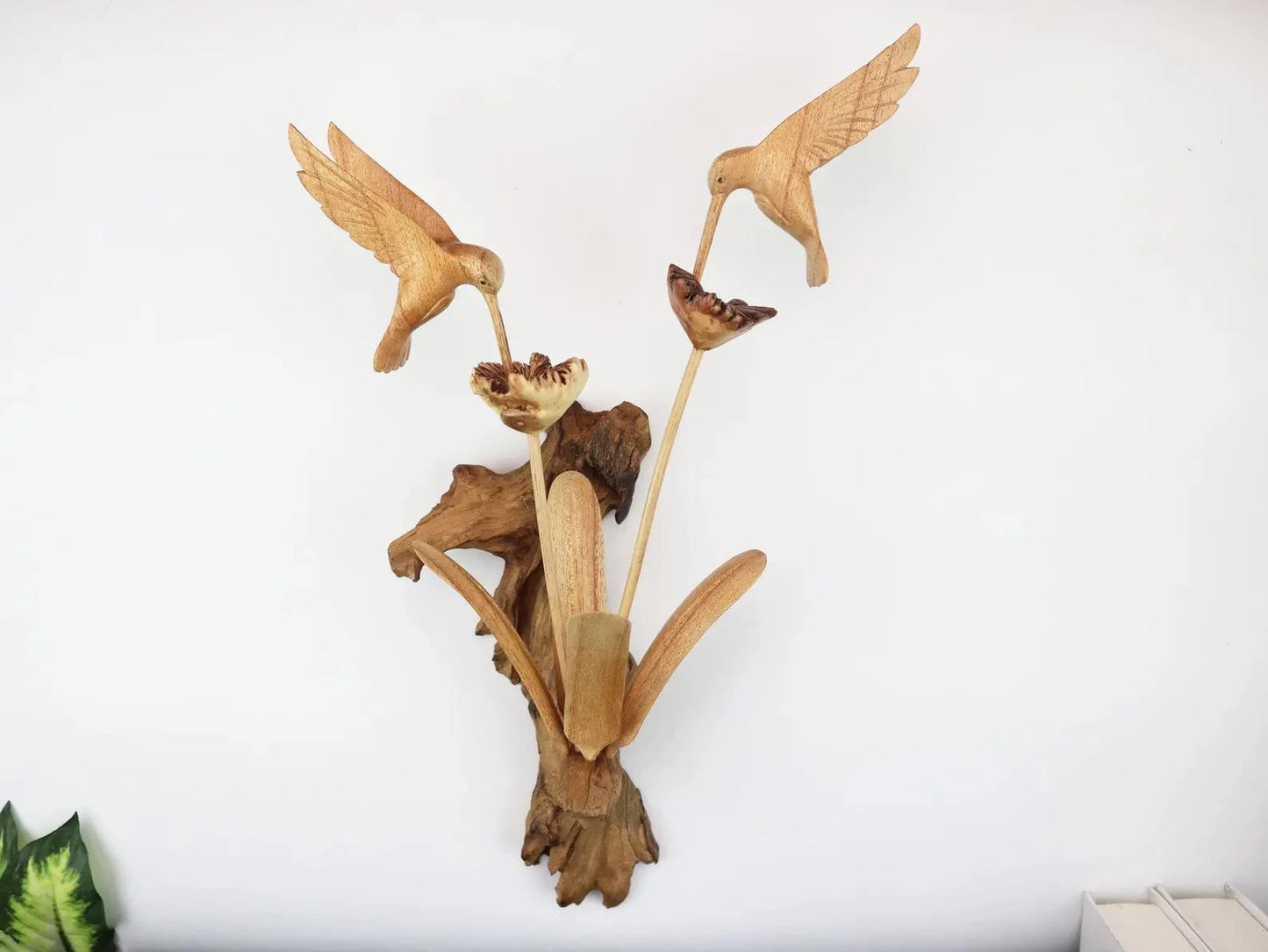 Hand-carved natural wooden wall art featuring a pair of hummingbirds in flight over two flowers, with an upright wood log or root base, rustic bird decor.
