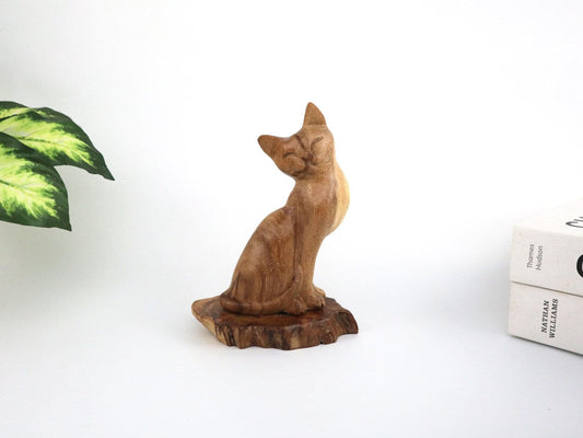 Hand-carved natural wooden cat sculpture in a sitting pose with tilted head, on an organic wood base, minimalist pet decor.
