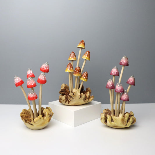 Set of three hand-carved wooden mushroom sculptures with slender stems and hand-painted caps in red, yellow, and purple colors on a rustic wood base, whimsical tabletop decor.