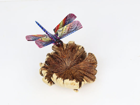 Hand-carved and painted wooden sculpture featuring a vibrant, multi-colored dragonfly on a textured pine cone, mounted on a round freestanding wooden base, rustic insect decor.