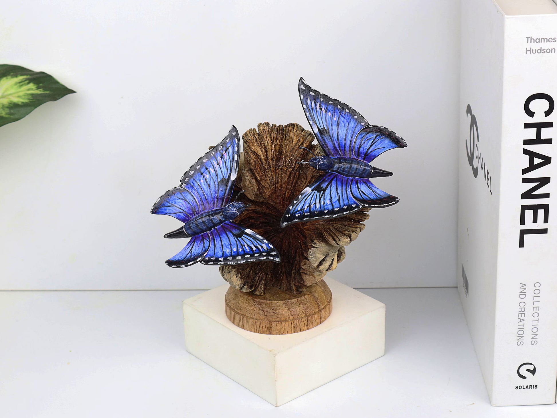 Hand-carved and painted wooden sculpture featuring two vibrant blue butterflies on a gnarled upright wood branch, mounted on a round freestanding wooden base, rustic decor.